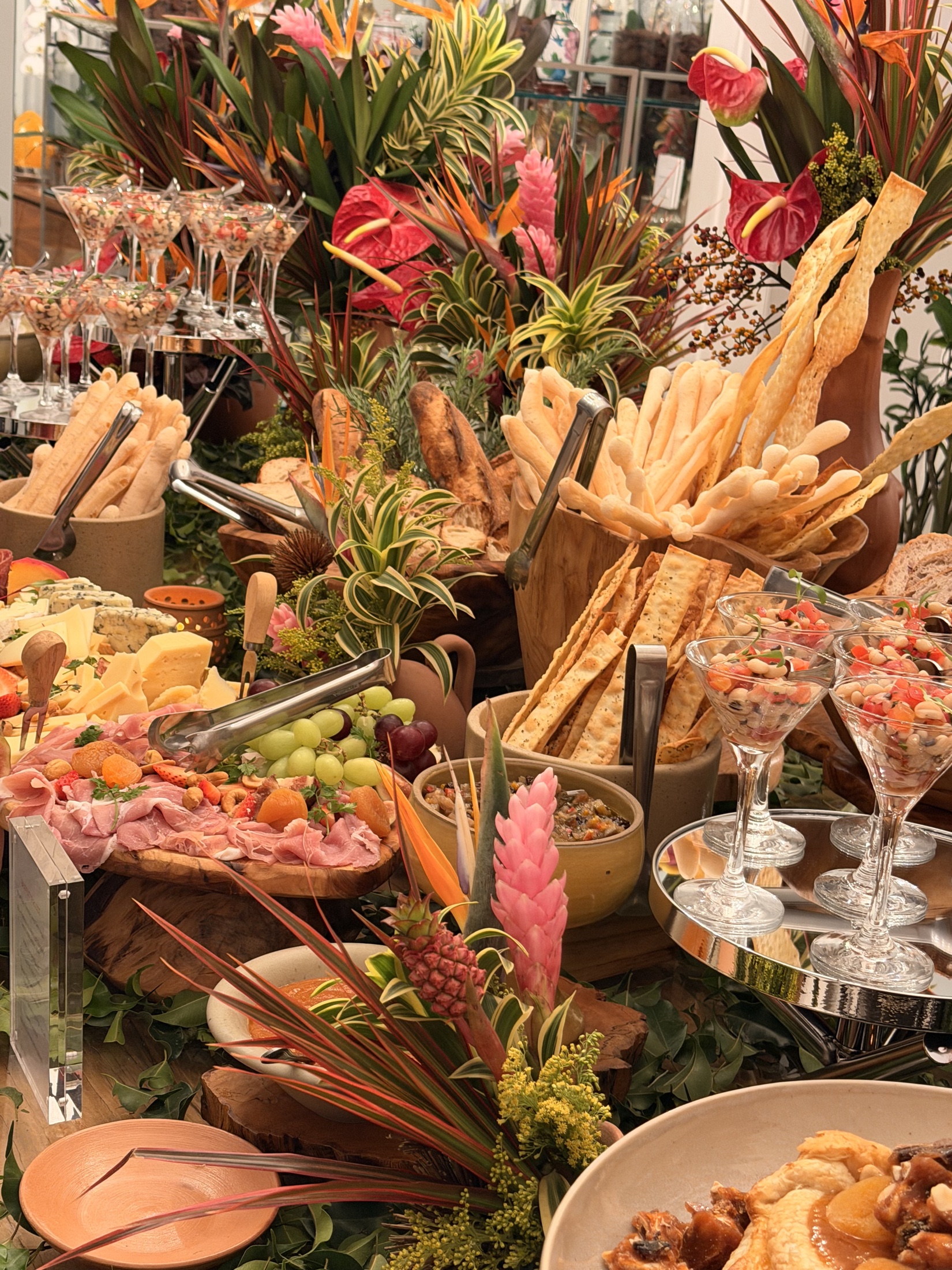 Mesa de buffet com pães, queijos, canapés e arranjos tropicais